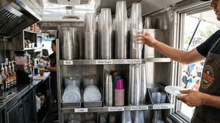 Food truck interior with organized cups, lids, and beverage service, worker reaching for cup