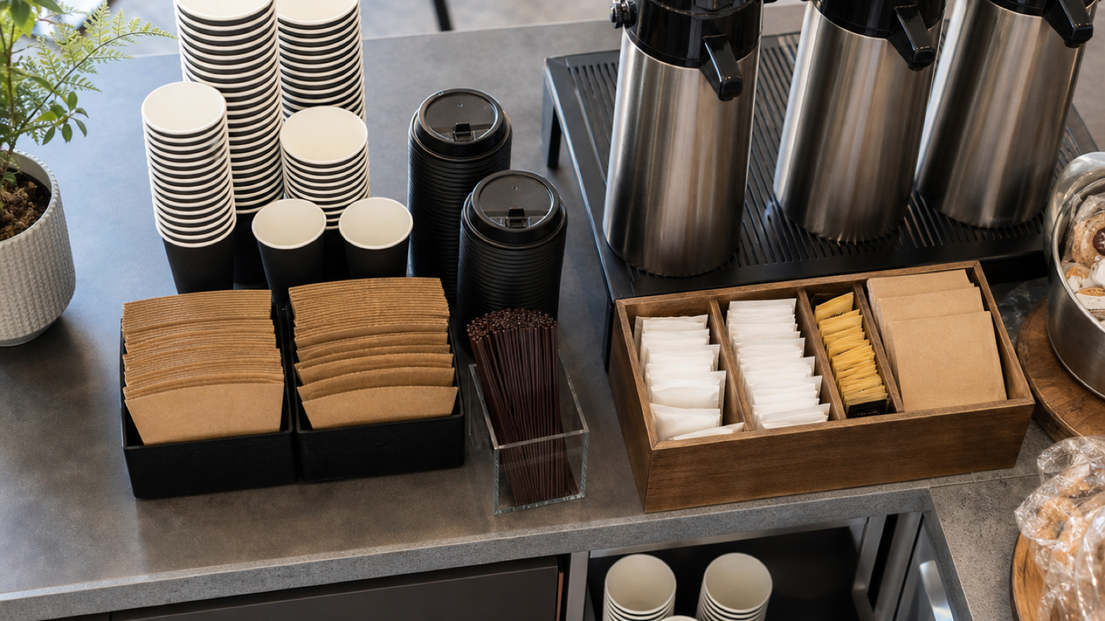 Professional café coffee station with black paper cups, lids, sleeves, airpots, and organized condiment zones