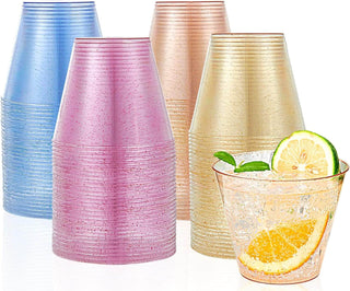 Colorful plastic cups in blue, pink, gold, and a clear glass with a fizzy drink, lemon, and mint on a reflective surface.