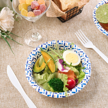 A colorful salad with lettuce, cherry tomatoes, egg, and pumpkin in a patterned paper bowl, accompanied by candy and bread.