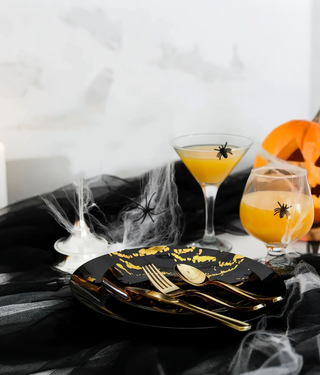 Halloween table setting with black plates, gold cutlery, spooky drinks, spiderwebs, and a jack-o'-lantern.