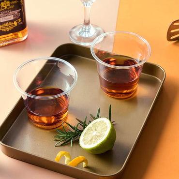 Two clear cups with dark brown drinks sit on a gold tray, alongside fresh lemon and rosemary garnishes. A whiskey bottle is visible.