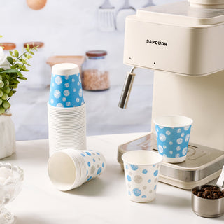 A stack of blue-patterned JOLLY CHEF 5 oz Paper Cups beside a coffee machine, with a few cups on the counter and coffee beans in a metal container.