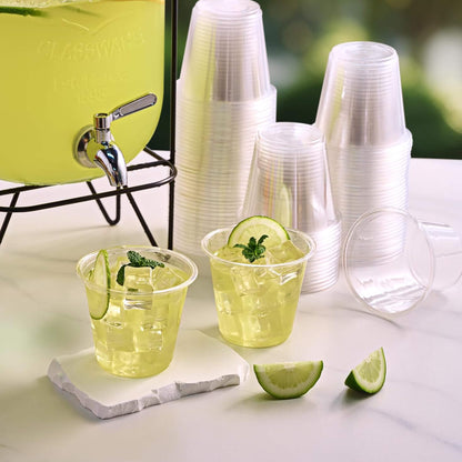 Two cups of iced green lemonade with mint and lime slices sit beside a drink dispenser and stacks of clear cups.