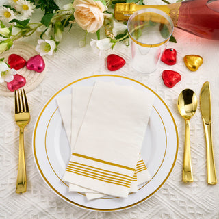 3 pieces of Jolly Chef 3 Ply Golden Stripe Classic Paper Napkins are placed on the white golden rimmed plate with spoon, fork and knife beside it on the white tablecloth