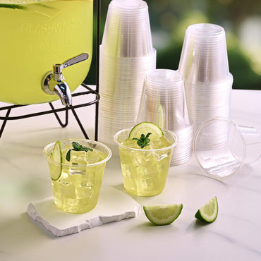 Two clear cups with green beverage, ice, mint, and lime slices are beside a drink dispenser and stacked plastic cups on a white surface.
