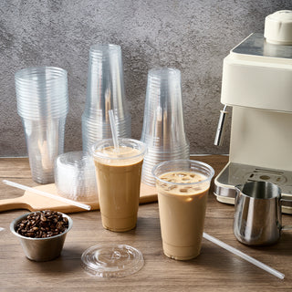 Two iced coffee drinks sit next to stacks of plastic cups, a coffee machine, and a bowl of coffee beans on a wooden countertop.