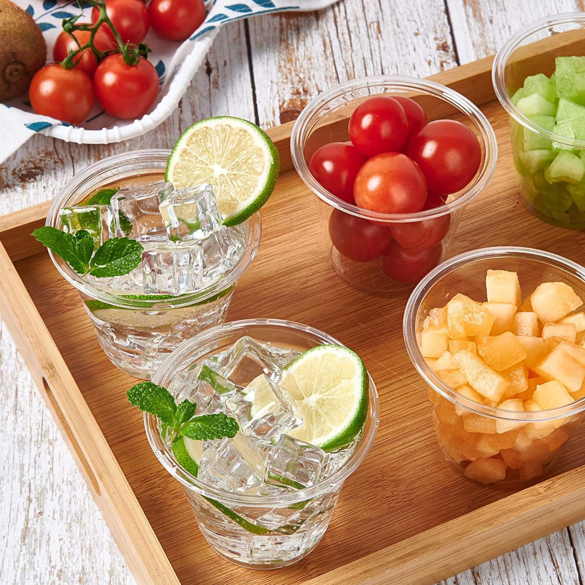 Two refreshing drinks with ice, mint, and lime slices, alongside cups of cherry tomatoes and diced cantaloupe, on a wooden tray.