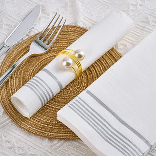 1 piece of 3 Ply Silver Pattern Classic Paper Napkin is wrapped with golden jewelry strap and placed in the middle of stack of napkin and fork, knife set on the white tablecloth.