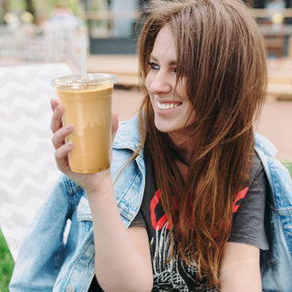 A person in a denim jacket holds a transparent cup of iced coffee, enjoying a relaxed outdoor setting.