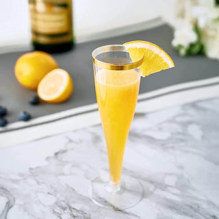 Jolly Chef 4.5oz Clear Gold Rim Champagne Flutes,  filled with a bright yellow cocktail, garnished with a lemon slice, beside whole and sliced lemons and blueberries.