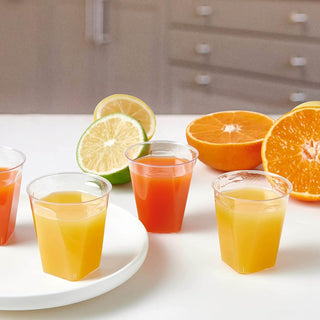 A variety of colorful fruit juices in Jolly Chef clear cups, with fresh oranges and limes cut in half nearby, all set on a white table.