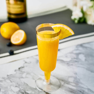 A vibrant orange drink in a Jolly Chef 5oz Clear Gold Rim Easy Grip Champagne Flutes adorned with a lemon slice, set on a marble table with lemons and greenery in the background.
