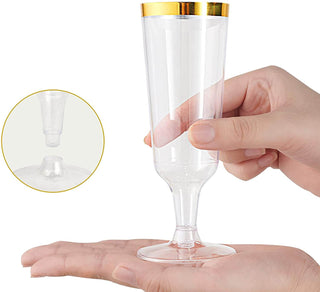 A person's hand holds a Jolly Chef 5oz Clear Gold Rim Easy Grip Champagne Flutes with a gold rim, demonstrating its unique pour spout feature.