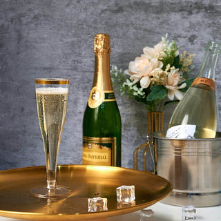 Jolly Chef 4.5oz Clear Gold Rim Champagne Flutes filled with bubbly sits on a gold tray, beside a green champagne bottle, ice cubes, and a bouquet of flowers.