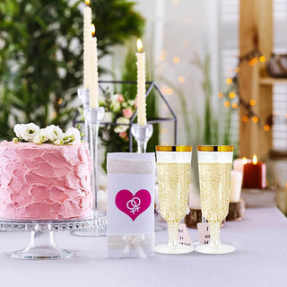A pink cake, Jolly Chef 5oz Clear Gold Rim Easy Grip Champagne Flutes, and candles create a romantic setting, featuring a gift wrapped in white with a pink heart symbol.
