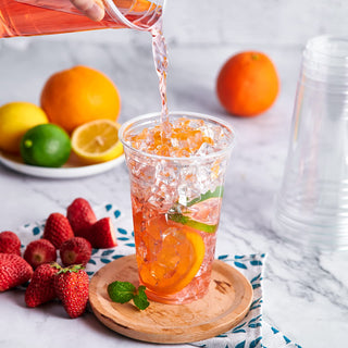 Clear PET plastic cup receiving pink drink with ice, orange, lime slices. Strawberries & citrus fruits nearby.