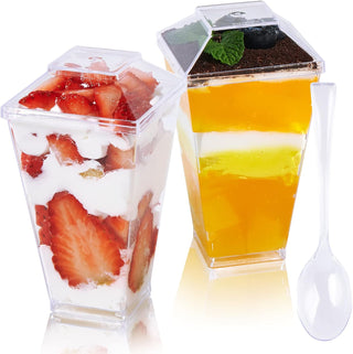 Two transparent cups contain layered desserts: one with strawberries and cream, the other with colorful jelly and whipped topping.