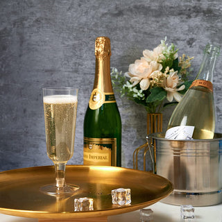 A golden tray with Jolly Chef 5oz Clear Easy Grip Champagne Flutes, two bottles of champagne, ice cubes, and a bouquet of flowers against a grey background.