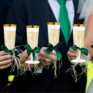 Four elegantly decorated Jolly Chef 5oz Clear Gold Rim Easy Grip Champagne Flutes with green bows, held by wedding guests, filled with bubbly beverage in bright sunlight.