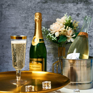 A sparkling Jolly Chef 5oz Clear Gold Rim Easy Grip Champagne Flutes
on a gold tray, surrounded by bottles, ice cubes, and a floral arrangement, set against a textured backdrop.