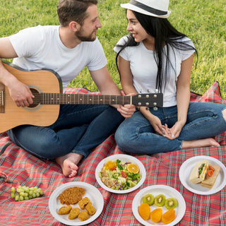 A couple enjoys a picnic on a plaid blanket, surrounded by jolly chef plates of food, while one plays guitar and the other listens.