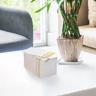 A stack of Jolly Chef 3 Ply Golden Stripe Classic Paper Napkins is placed on the white table next to the green plant in the living room with sofa next to the table