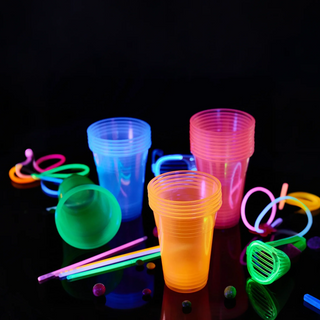 Colorful glow sticks, cups, and glasses on a black reflective surface. Party supplies glowing.