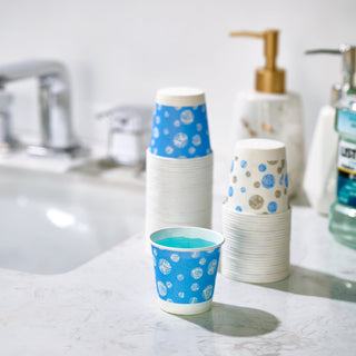 A stack of JOLLY CHEF 3 oz Paper Cups – 300 Count, Small Bathroom Cups, Disposable Mouthwash Cups, Espresso Cups, Mini Cup for Coffee, Tea, Water, Snack in blue and white sits on a sink, with a faucet and bathroom products visible in the background.