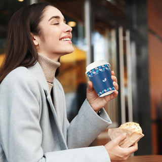 A person in a gray coat and turtleneck holds Jolly Chef Blue&Grey Leaf Pattern Coffee Cups With Lids&Sleeves & Stirrers in one hand and a pastry wrapped in paper in the other.