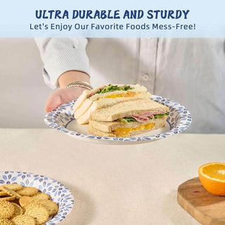 A person holds a sturdy paper jolly chef plate with layered sandwiches, accompanied by a plate of cookies and a halved orange on a table.
