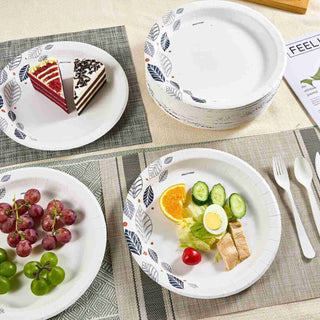 A table setting features colorful jolly chef plates of grapes, salad, and two cake slices, with stacked white paper plates and utensils on a textured tablecloth.