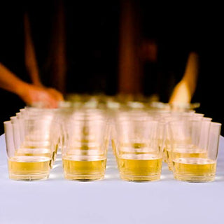 A row of clear glasses filled with golden liquid, with a blurred hand in the background, suggesting a social gathering or tasting event.